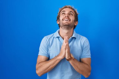 Beyaz bir adam mavi arka planda duruyor. Elleriyle yalvarıyor ve dua ediyor. Yüzünde umut ifadesi var. Yalvarıyor. 