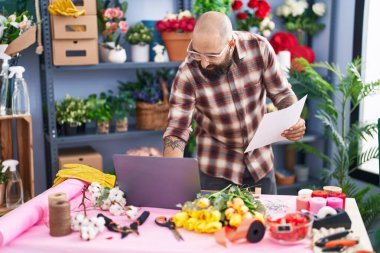 Young bald man florist using laptop reading document at flower shop