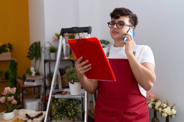 Non binary man florist talking on smartphone reading clipboard at flower shop