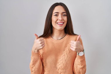 Beyaz arka plan başarı işaretinin üzerinde duran genç esmer kadın elleriyle olumlu jest yapıyor, gülümsüyor ve mutlu. Neşeli ifade ve kazanan jest. 