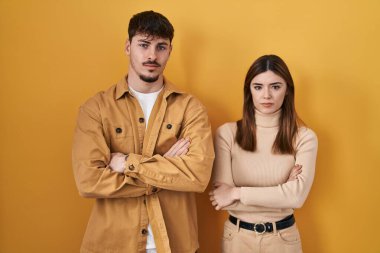 Sarı arka planda duran genç İspanyol çift şüpheci ve gergin, kollarını çapraz yapıp yüz ifadesini onaylamıyorlar. negatif kişi. 