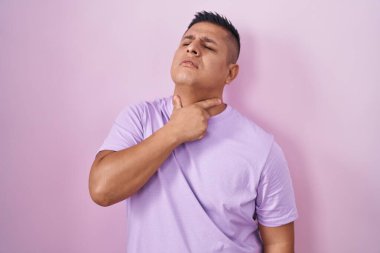 Young hispanic man standing over pink background touching painful neck, sore throat for flu, clod and infection 