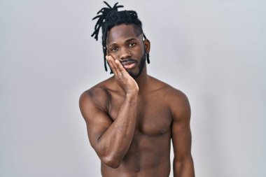 Afrikalı bir adam, gömleksiz bir şekilde arka planda duruyor. Kollarını kavuşturup depresyondan sıkılmış ve yorgun görünüyor.. 