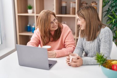 Anne ve kızı evde dizüstü bilgisayar kullanarak kahve içiyorlar.