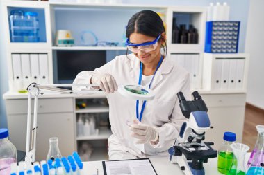 Laboratuvarda büyüteç kullanan genç Latin kadın bilim adamı üniforması giyiyor.