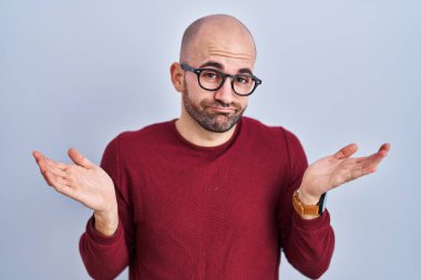 Young bald man with beard standing over white background wearing glasses clueless and confused expression with arms and hands raised. doubt concept.  clipart