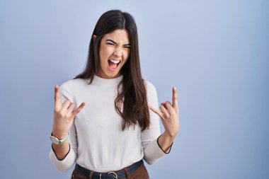 Genç esmer kadın mavi arka planda duruyor çılgın bir ifadeyle bağırıyor elleri havada rock sembolü yapıyor. Müzik yıldızı. ağır müzik konsepti. 