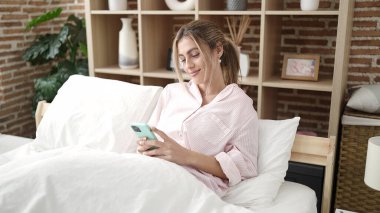 Yatak odasında akıllı telefon kullanan genç sarışın kadın.