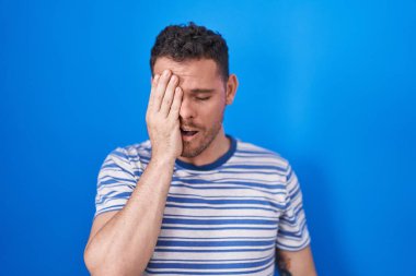 Young hispanic man standing over blue background yawning tired covering half face, eye and mouth with hand. face hurts in pain. 