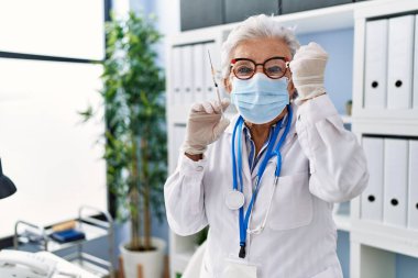 Gri saçlı, doktor üniforması giyen ve elinde şırınga tutan yaşlı bir kadın öfkeyle bağırıyor, öfkeyle bağırıyor ve el kaldırıyor. 