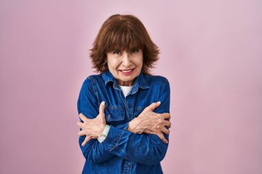 Middle age woman standing over pink background shaking and freezing for winter cold with sad and shock expression on face 