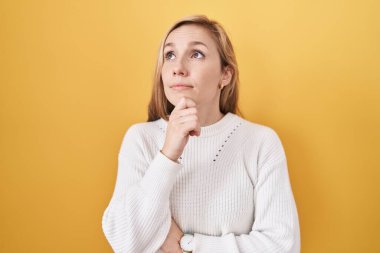 Sarı arka plan üzerine beyaz kazak giyen, eli çenesinde soru soran beyaz bir kadın, düşünceli bir ifade. Düşünceli bir yüzle gülümsüyor. şüphe kavramı. 