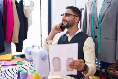 Genç Arap terzi akıllı telefondan konuşuyor giyim fabrikasında kıyafet tasarımı yapıyor.