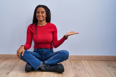 Saçları örgülü genç Afro-Amerikalı evde oturmuş gülümsüyor ve avuç içi kameraya bakıyor.. 