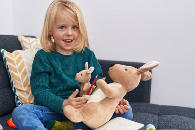 Evdeki kanepede oturan, elinde renkli kalemler tutan sevimli beyaz çocuk.