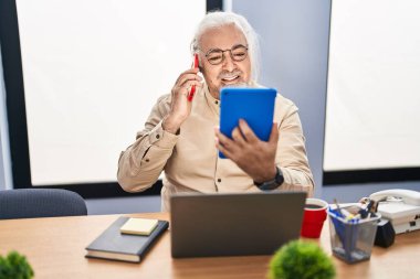 Middle age grey-haired man business worker talking on smartphone using touchpad at office