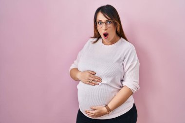 Pembe arka planda duran hamile bir kadın şaşkınlık ve şaşkınlık dolu bir ifade, korku ve heyecan dolu bir yüz.. 
