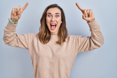 Young woman standing over isolated background smiling amazed and surprised and pointing up with fingers and raised arms. 