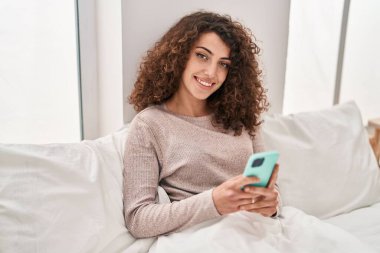 Young hispanic woman using smartphone sitting on bed at bedroom