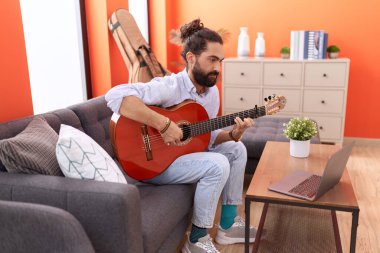 Young hispanic man having online guitar class sitting on sofa at home