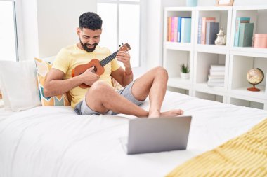 Young arab man having online ukulele lesson sitting on bed at bedroom