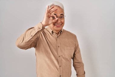 Hispanic senior man wearing glasses doing ok gesture with hand smiling, eye looking through fingers with happy face. 