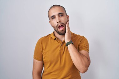 Hispanic man with beard standing over white background touching mouth with hand with painful expression because of toothache or dental illness on teeth. dentist 
