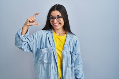 Mavi arka planda duran, gülümseyen, kendine güvenen, el hareketi yapan, parmakları küçük boy işareti yapan ve kameraya bakan genç İspanyol kadın. ölçüm kavramı. 