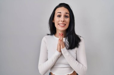 Hispanik bir kadın tecrit edilmiş arka planda duruyor ellerini birleştirip af diliyor kendinden emin bir şekilde gülümsüyor. 