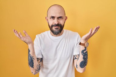 Young hispanic man with tattoos standing over yellow background clueless and confused expression with arms and hands raised. doubt concept. 