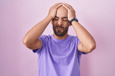 Orta yaşlı kel bir adam pembe arka planda dikiliyor. Baş ağrısı, çaresiz ve stresli. Ağrı ve migren yüzünden. Eller başının üstünde.. 