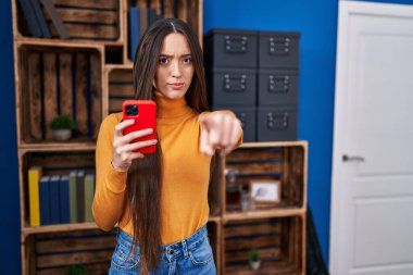 Genç esmer kadın kamerayı parmağıyla işaret ederek akıllı telefonu kullanıyor ve sen kendine güvenen ciddi bir hareket yapıyorsun. 