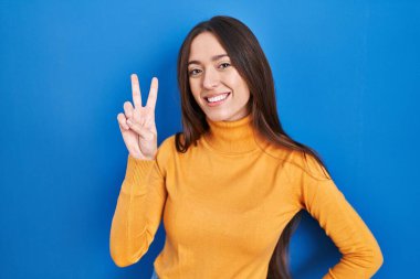 Mavi arka planda duran esmer kadın kameraya bakıp zafer işareti yapıyor. İki numara.. 