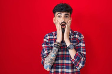 Kırmızı arka plan üzerinde duran sakallı genç İspanyol adam korkmuş ve şok olmuş, şaşırmış ve şaşırmış bir yüz ifadesi var. 