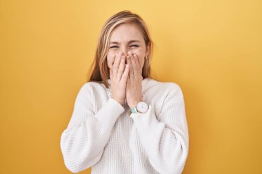 Sarı arka plan üzerine beyaz kazak giyen genç beyaz kadın gülüyor ve utanıyor kıkırdıyor ağızlarını ellerle, dedikoduyla ve skandal konseptiyle kapatıyor. 