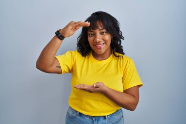 Hispanic woman standing over blue background gesturing with hands showing big and large size sign, measure symbol. smiling looking at the camera. measuring concept. 