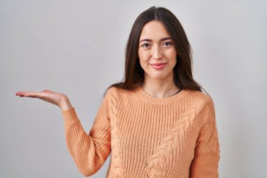 Genç esmer kadın beyaz arka planda duruyor. Gülümsüyor, mutlu bir şekilde sunuyor ve avuç içi kameraya bakıyor.. 