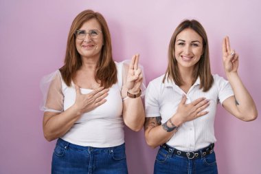 İspanyol anne ve kızı pembe arka planda rahat beyaz t-shirt giyiyorlar gülümsüyorlar, elleri göğsünde ve parmakları havada yemin ediyorlar, sadakat yemini ediyorlar. 