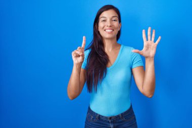 Mavi arka planda dikilen genç İspanyol kadın altı numaralı parmaklarını gösterip işaret ederken kendinden emin ve mutlu gülümsüyor.. 