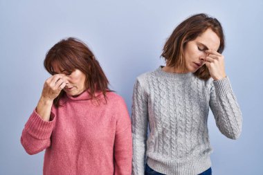 Anne ve kızı mavi arka planda dikiliyorlar yorgun burunlarını ve gözlerini ovuşturuyorlar yorgunluk ve baş ağrısı hissediyorlar. Stres ve hüsran kavramı. 