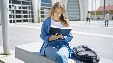 Üniversitede bankta oturmuş kitap okuyan genç sarışın kadın.