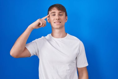 Kafkasyalı sarışın adam mavi arka planda durup gülümsüyor. Tek parmakla işaret ediyor. Harika bir fikir ya da düşünce, iyi bir hafıza. 