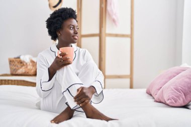 Young african american woman drinking cup of coffee sitting on bed at bedroom