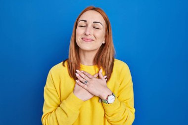Young woman standing over blue background smiling with hands on chest with closed eyes and grateful gesture on face. health concept. 