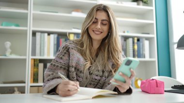 Young blonde woman student using smartphone writing on notebook at library university