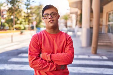 Down sendromlu adam gülümsüyor. Kollarını kavuşturup caddede duruyor.