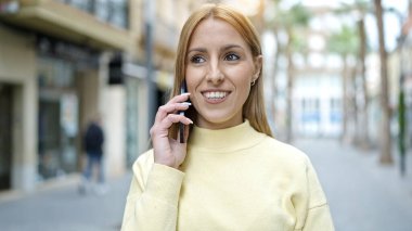 Genç sarışın kadın gülümsüyor. Kendine güveni tam. Akıllı telefondan konuşuyor.