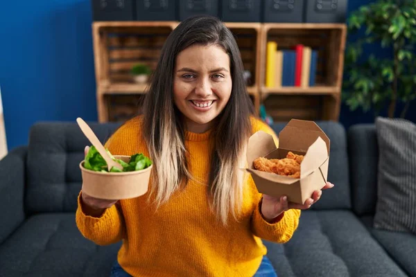 stock image Young hispanic woman holding healthy salad and fried chicken wings smiling with a happy and cool smile on face. showing teeth. 