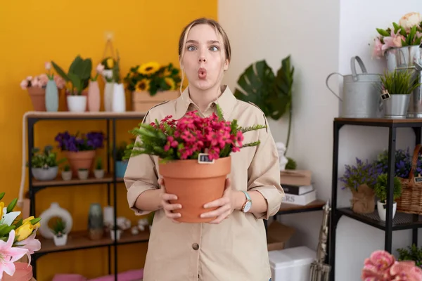 Young caucasian woman working at florist shop holding plant pot making fish face with mouth and squinting eyes, crazy and comical.