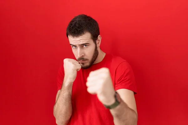 若いヒスパニック系の男 怒りと動揺の顔 問題を恐れて 拳の防衛ジェスチャーで戦うために準備ができてカジュアルな赤いTシャツを着て — ストック写真